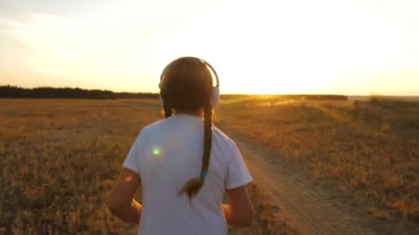 Muchacha joven en blanco T-shirt entrena al atardecer y escucha música con auriculares. haciendo footing deportivo al atardecer fuera de la ciudad — Vídeo de stock