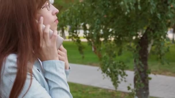 Femme d'affaires dans des lunettes parler sur smartphone rire et boire du café tout en marchant dans le parc. gros plan — Video