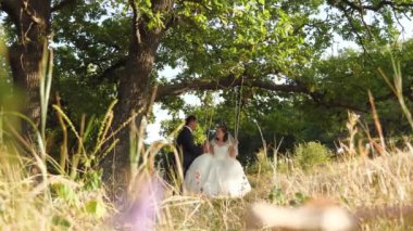 Gelin ve damat yaz parkta salıncak üzerinde sallanan güzel beyaz elbiseli. Yaz ormandaki bir meşe dalı üzerine salıncak.