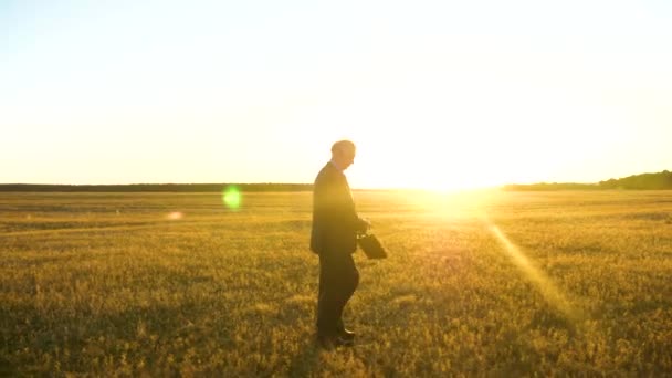 Man in donker pak met zwarte aktetas wandelen door veld in stralen van een stralende zon voor een belangrijke vergadering — Stockvideo