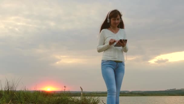 Meisje in stralen van zonsondergang wandelen op strand en controleren van mail op een tablet online. meisje met koptelefoon wandelen langs strand met Tablet PC- en luisteren naar muziek. — Stockvideo