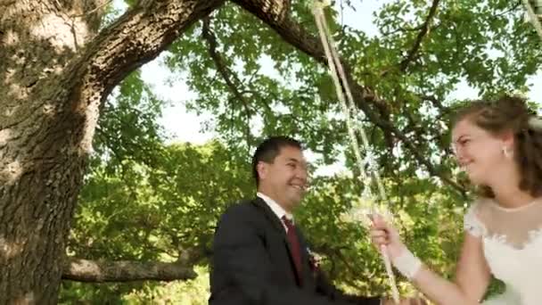 Gelukkige bruid en bruidegom in prachtige witte jurk swingen op schommel in zomer park. Swing op tak van een eik in zomer bos. — Stockvideo