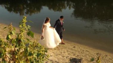 beyaz elbise ve damat takım elbise yalınayak yürümek nehir kenarı boyunca kum üzerinde mutlu gelin. seven çift sahilde elinde el gidiyor.