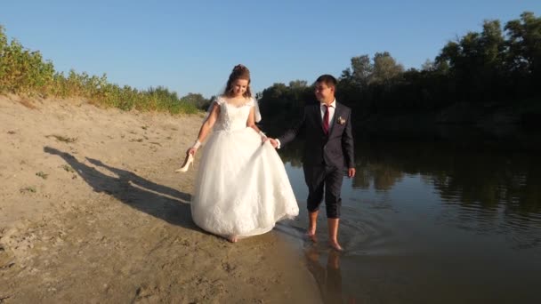 Όμορφη νύφη σε λευκό φόρεμα και γαμπρός περπατάτε ξυπόλητοι στο νερό κατά μήκος της όχθης του ποταμού. αγάπη ζευγάρι πηγαίνει χέρι-χέρι στην παραλία. — Αρχείο Βίντεο