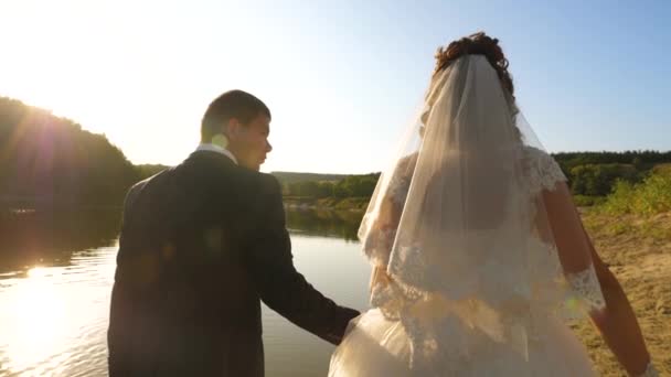 Bride in white dress and groom walk barefoot along edge of river bank. loving couple goes hand in hand on the beach. Slow motion — Stock Video