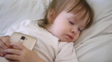 Smartphone ile yatakta uyuyan sevimli bebek. Çocuk yastığın üzerinde yalan ve bir tablet holding