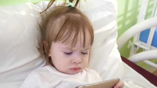 Roztomilá holčička leží na posteli v dětské nemocnici, sledovat vtipné karikatury na smartphone. — Stock video