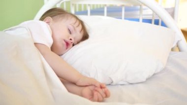 sevimli bebek beyaz yatak odasında evde onun yatakta uyuya kalır. Uyuyan Çocuk kavramı. Çocuk uyumak istiyor ve elleriyle gözlerini ovuyor. çocuk hastane koğuşunda uyur. yakın çekim