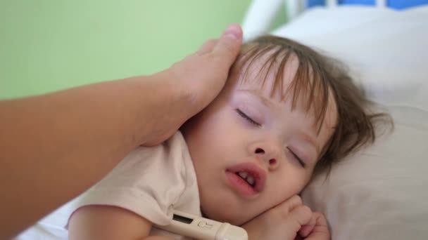 Mutter misst Temperatur mit Thermometer an Baby und streichelt Kind mit der Hand. Nahaufnahme. Baby schläft auf Krankenhausstation auf weißem Bett. — Stockvideo