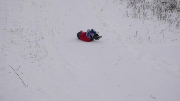 穿着红色夹克的快乐女孩在雪地里雪橇滚下山。快乐的孩子在圣诞公园玩冬天。慢动作 — 图库视频影像