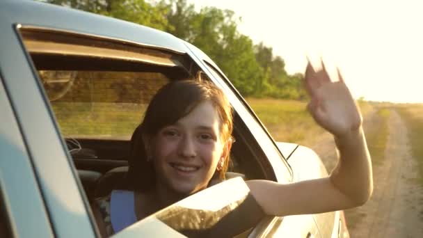 Bella ragazza con i capelli lunghi cavalca una macchina con le mani al finestrino e sorride agitando la mano nel bagliore del sole dorato. Rallentatore . — Video Stock