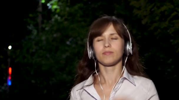 Joven hermosa mujer bailando, sonriendo y disfrutando de la música por la noche en el parque bajo la luz de las linternas. Primer plano. chica feliz escuchando música en los auriculares en el teléfono inteligente . — Vídeos de Stock