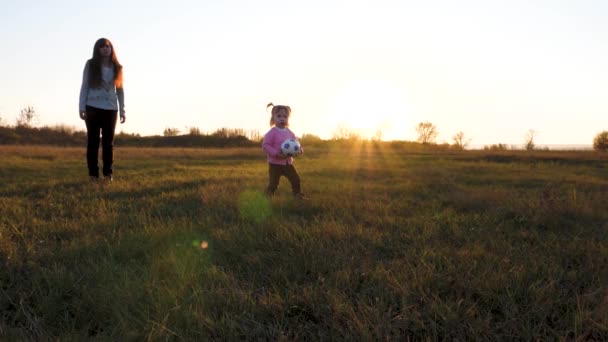 Criança feliz corre em campo com bola em suas mãos e joga na grama, mãe cuida de sua filha. O miúdo joga com a bola de futebol infantil no parque ao pôr-do-sol. Movimento lento — Vídeo de Stock