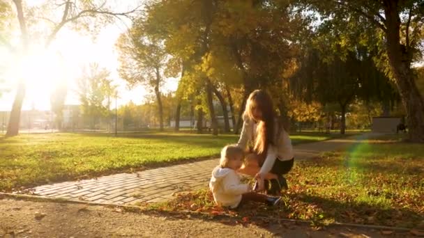 Mamãe brinca com o bebê com folhas amarelas secas no gramado no parque de outono. criança está sentada na grama, mamãe pega criança do gramado . — Vídeo de Stock