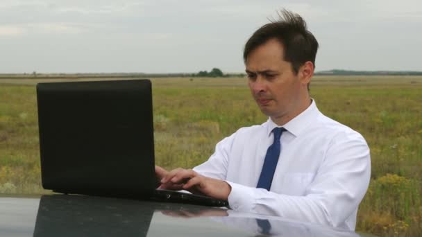 Male businessman in blue and white shirt is working in field on computer on roof of the car. — Stock Video