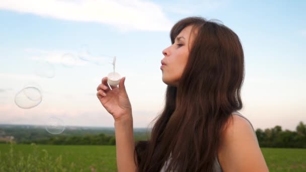 Beautiful girl with long hair blowing transparent soap bubbles against the blue sky and smiling.Slow motion. — Stock Video