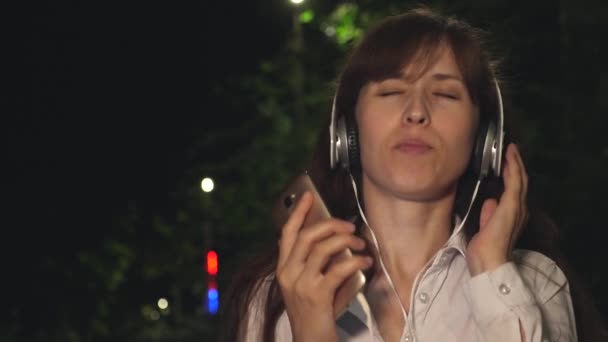 Flicka i hörlurar och touch telefon vifta med långt hår och dansande leende och får buzz från musiken på natten i park — Stockvideo