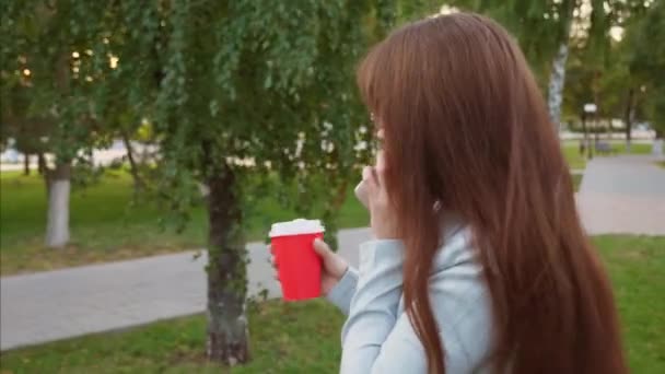 Felice donna d'affari con gli occhiali in tuta leggera cammina nel parco bevendo caffè e parlando su uno smartphone, ragazza ride — Video Stock