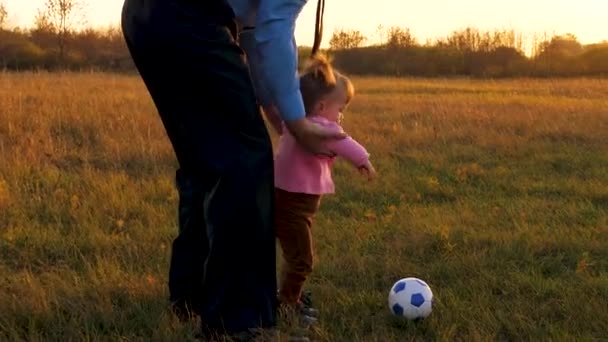 Ayah bahagia dan anak menendang bola. Keluarga bermain dengan bayi yang lebih muda dengan bola sepak di taman saat matahari terbenam. Konsep keluarga bahagia. Gerakan lambat — Stok Video