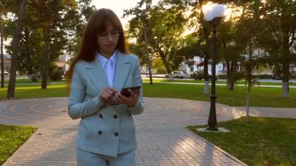 Mooie zakenvrouw wandelingen langs steegje in park met tablet in haar handen, meisje in een lichte pak gaat aan het werk. — Stockvideo