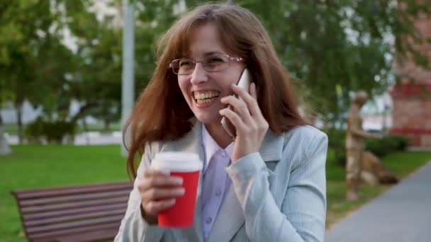 Hermosa chica está bebiendo café y hablando en el teléfono inteligente, chica se ríe. joven mujer en gafas en traje de luz va en el parque en verano — Vídeo de stock