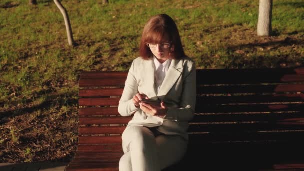 Beautiful female businessman wearing glasses and working with tablet and checks email in summer park on a bench. — Stock Video