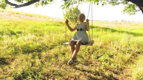 Chica feliz con el pelo largo se balancea en columpio bajo el roble de verano en un vestido blanco y se ríe . — Vídeo de stock