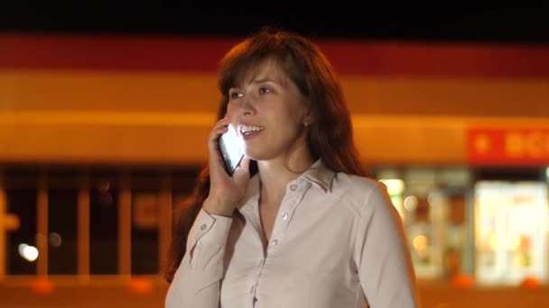 Young woman talking on the smartphone in parking lot at night by light of lanterns. Beautiful girl talking on phone at night in the city. — Stock Video
