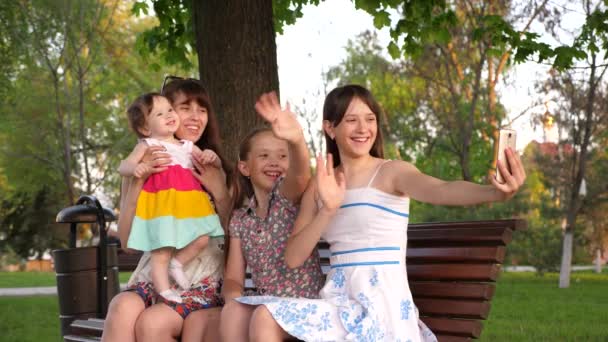 Joven madre de niños felices siendo fotografiada en un teléfono inteligente. una familia feliz sentada en el banco en el parque haciendo salphy en el teléfono sonriendo y agitando sus manos . — Vídeos de Stock