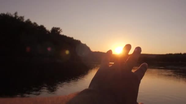 남자의 손은 태양에 대 한에 도달합니다. 햇빛은 손가락을 통해 빛난다. 아름 다운 zakt 여름 강입니다. 슬로우 모션 — 비디오
