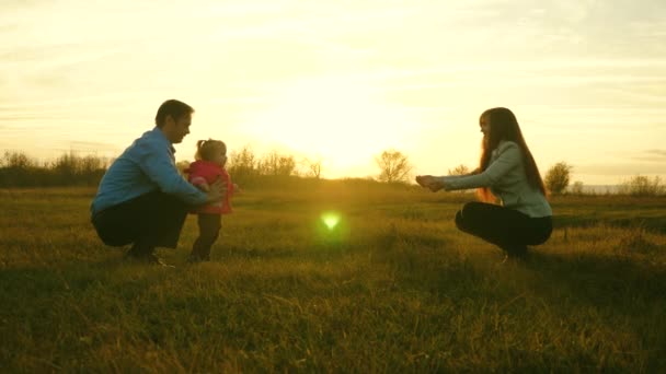 Дитина йде на газон від тата до мами. дитина робить перші кроки в парку. мама і тато грають з дитиною на траві на заході сонця. концепція сімейного щастя — стокове відео