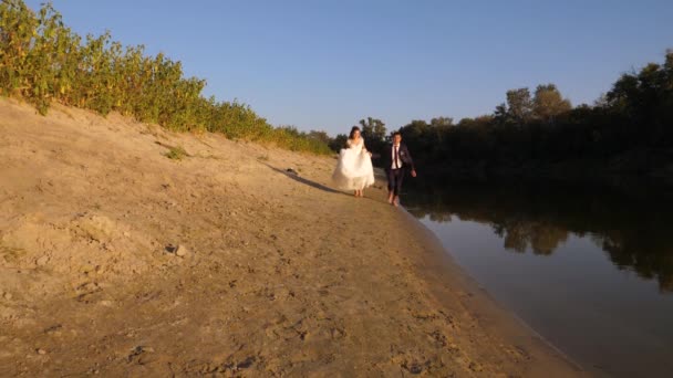 Couple amoureux court le long du bord de la rivière. marié heureux en costume et mariée en robe blanche courir sur la plage. concept d'une famille heureuse . — Video