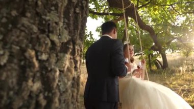 Parkta ağaç üzerine mutlu gelin ve damat ipte sallanan salıncak. aşk kaç içki şarap içinde birbirlerine gülümseyen güzel gözlük. mutlu aile kavramı.