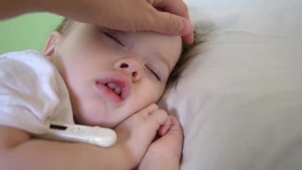 Child is sick and sleeps with thermometer on white bed. Mom measures temperature with thermometer for the baby and caresses kid with her hand. close-up. — Stock Video