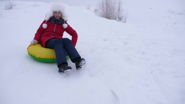 Счастливые детские слайды на снежных горках на санях, трубах. Девушка играет зимой в парке. веселая девушка-подросток в белой шляпе и красной куртке смеется — стоковое видео