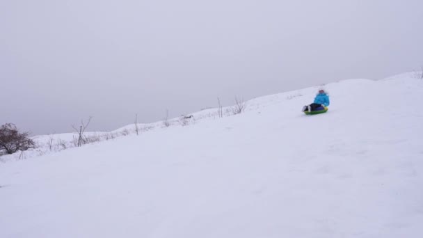 Bela jovem desliza para baixo slide na neve em um tubo de neve inflável e acena sua mão. Menina feliz desliza através da neve no trenó. menina jogando no parque para as férias de Natal no inverno — Vídeo de Stock