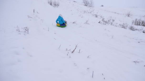 Счастливая девушка скользит по снегу по надувной снежной трубке и машет рукой. красивая молодая женщина скользит по снегу на санях. девушка играет в парке на рождественские каникулы зимой — стоковое видео