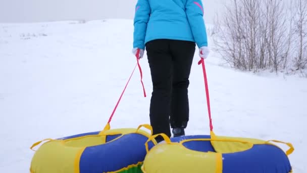 Chica sube montaña nevada y tira del trineo y un tubo de nieve inflable. descanso en el parque de invierno para las vacaciones de Navidad — Vídeos de Stock