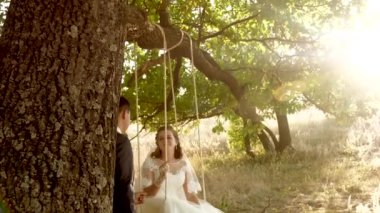 mutlu damat yaz parkta güzel beyaz elbiseli salıncak gelin üzerinde sallanan. Yaz ormandaki bir meşe ve sevgi dolu bir kaç dalı üzerinde salıncak