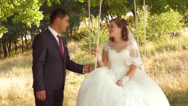 Happy bride and groom in beautiful white dress swinging on swing in summer park. swing on branch of an oak in summer forest and a loving couple — Stock Video