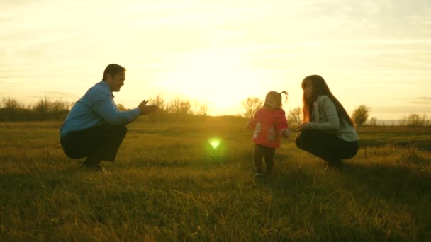 Dítě jde a spadá na trávě od mámy k tátovi. otec drží dcera dítě a polibky. dítě dělá své první krůčky. Koncept rodinného štěstí, krásné slunce, západ slunce. — Stock video