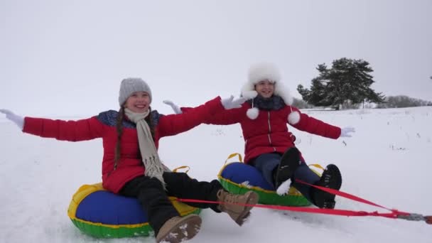 As crianças deslizam na neve em um tubo de neve inflável e ondas mão. Meninas felizes desliza através da neve no trenó. crianças brincando no parque para as férias de Natal no inverno — Vídeo de Stock