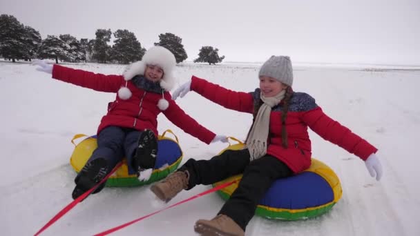 Glückliche Kinder rodeln im Winter im Schnee und winken mit den Händen. Kinder lachen und jubeln. Mädchen spielen in den Weihnachtsferien im Winterpark. Zeitlupe — Stockvideo