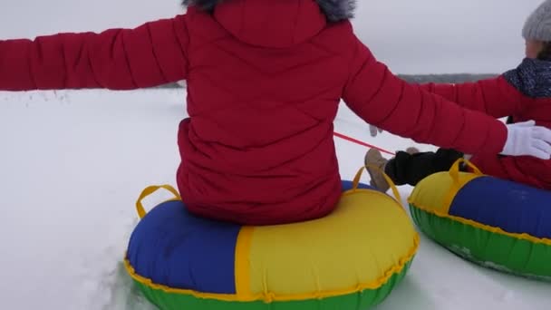 Kinderen van dia's in de sneeuw op een opblaasbare sneeuw tube en golven van de hand. kinderen spelen in het park voor de kerstvakantie in de winter. Gelukkig meisjes dia's door sneeuw op slee. — Stockvideo