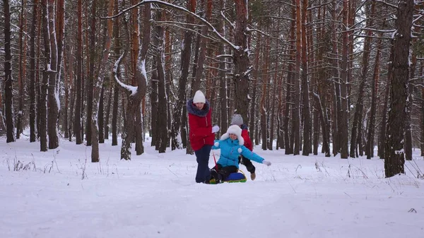 Lustige Kinder fahren mit ihrer Mutter auf Schlitten und einem aufblasbaren Schneeschlauch im Kiefernwald. glückliche Familie Mutti und Kinder spielen im Winter Park und Wald für die Weihnachtsfeiertage. — Stockfoto