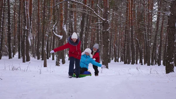 Lustige Kinder fahren mit ihrer Mutter auf Schlitten und einem aufblasbaren Schneeschlauch im Kiefernwald. glückliche Familie Mutti und Kinder spielen im Winter Park und Wald für die Weihnachtsfeiertage. — Stockfoto