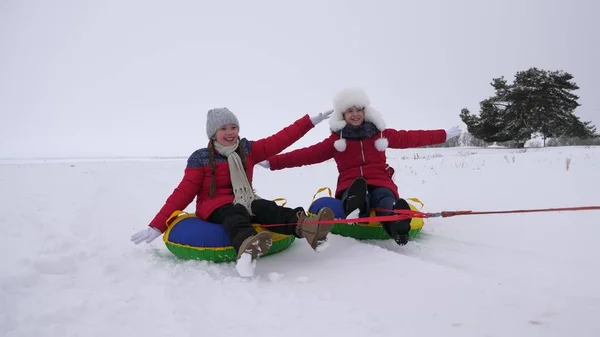 Barn glider i snön på en uppblåsbar snow tube och vågor hand. Glada tjejer bilder genom snön på släde. barn som leker i parken för julhelgen på vintern — Stockfoto