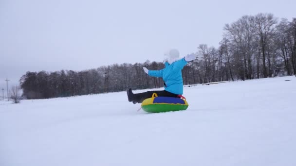 Niña se desliza hacia abajo se desliza en la nieve en un tubo de nieve inflable y agita su mano. Chica feliz jugando en el parque para las vacaciones de Navidad en invierno. Movimiento lento . — Vídeos de Stock