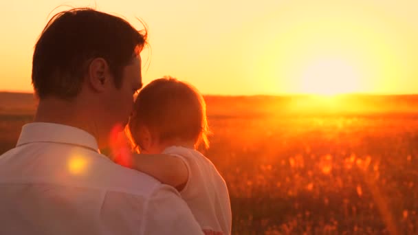 Papá lleva a su pequeña hija en sus brazos, iluminada por rayos de hermoso atardecer. Felices caminatas familiares en el parque en verano. Movimiento lento . — Vídeos de Stock