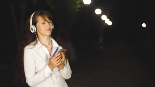 Mujer con auriculares escuchando bailes de canciones y canta hojeando a través del teléfono táctil en el parque nocturno de la ciudad — Vídeo de stock